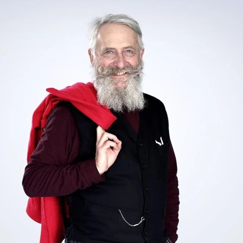 Un homme âgé avec une barbe grise, portant un pull noir et une veste marron, tient une veste rouge sur son épaule, souriant en regardant vers l'avant, posé contre un fond blanc.
