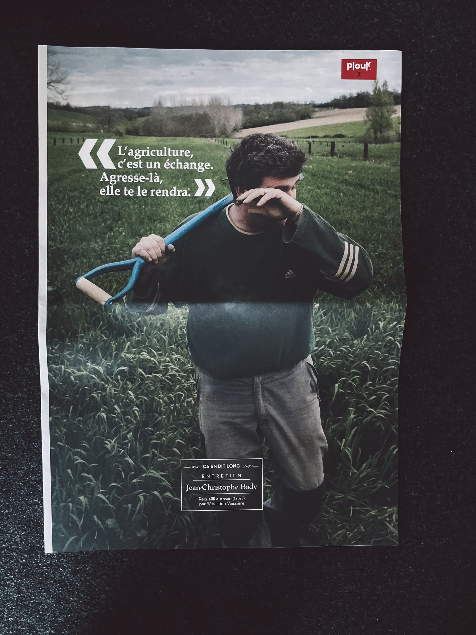 Affiche de magazine montrant un jeune homme marchant dans un champ avec une pioche sur l'épaule. Texte en français : "L'agriculture, c'est un échange. Agressé là, elle te le rendra."