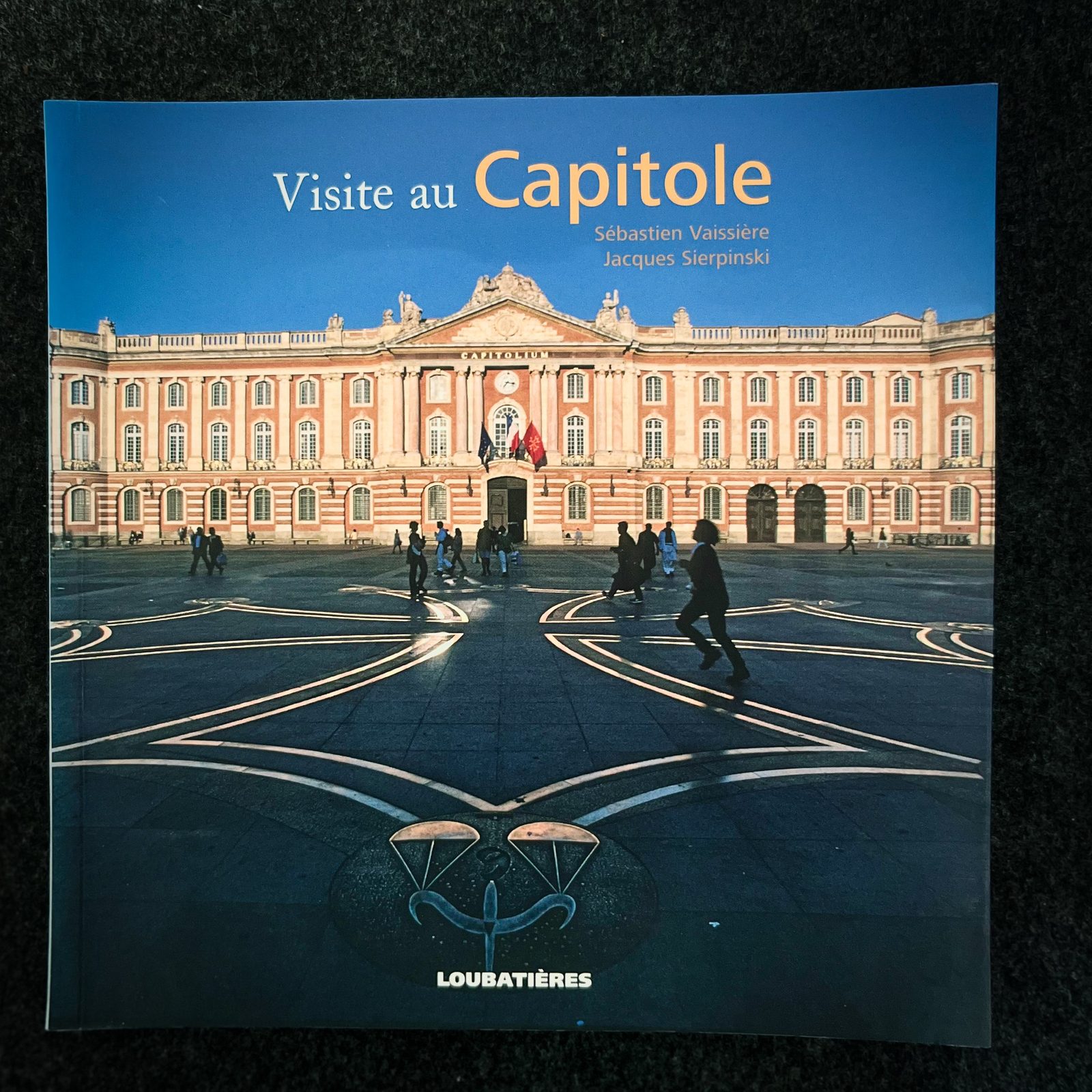Couverture d'un livre intitulé 'Visite au Capitole' avec une photo du Capitole de Loubatières, montrant un bâtiment historique en pierre rose avec des personnes marchant devant, au clair de la journée.